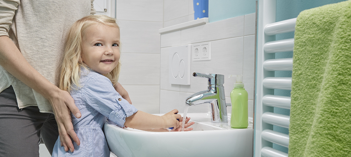 Ein blondes Mädchen in einem hellblauen Kleid wäscht sich die Hände am Waschbecken.