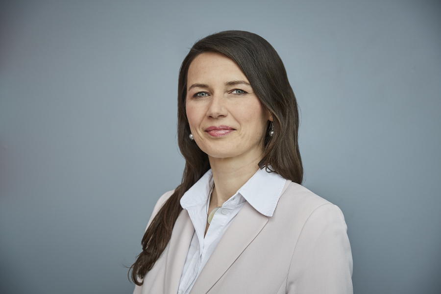 Das Bild zeigt eine Porträtaufnahme der Pressesprecherin Iris Rudolph. Sie hat lange braune Haare, die über eine Schulter fallen und lächelt in die Kamera. Sie trägt eine weiße Bluse und einen puderfarbenen Blazer. Der Hintergrund ist hellgrau.
