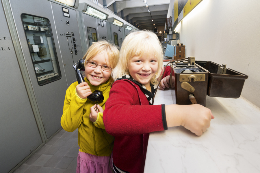 In der Bildmitte befinden sich zwei blonde Mädchen mit bunter Kleidung. Sie steht im Historischen Technikzentrum der Stadtwerke Halle und probieren die ausgestellten Exponate aus. Das linke Mädchen hält sich einen Telefonhörer an ihr Ohr und das Mädchen auf der rechten Seite bedient die Kurbel einer kleinen dunkelbraunen Box.